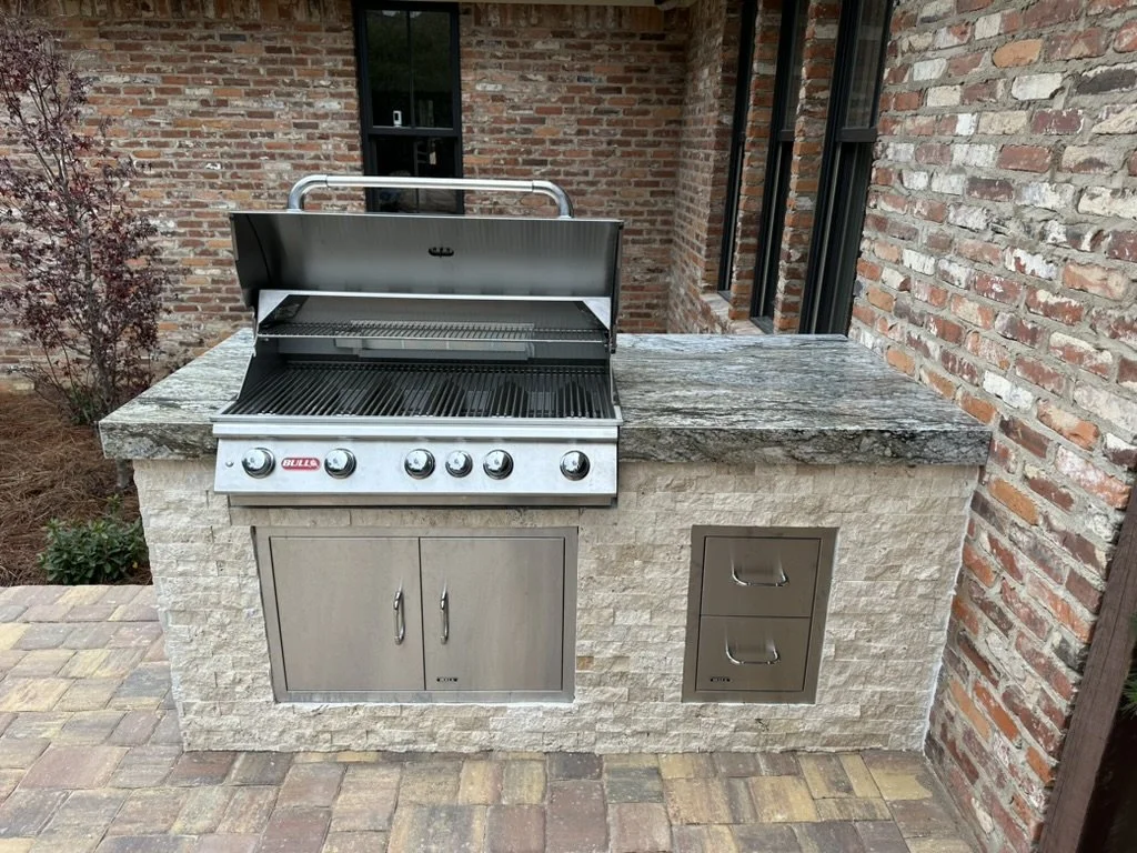 Outdoor stainless steel grill with a stone countertop and storage drawers on a brick patio, set against a rustic brick wall.