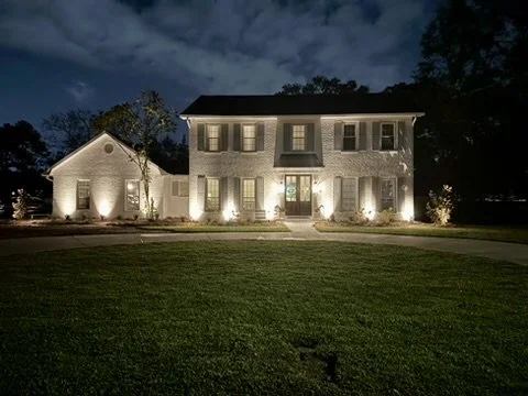Two-story white brick house illuminated by landscape lighting, with a circular driveway and well-maintained front yard at night under a cloudy sky.