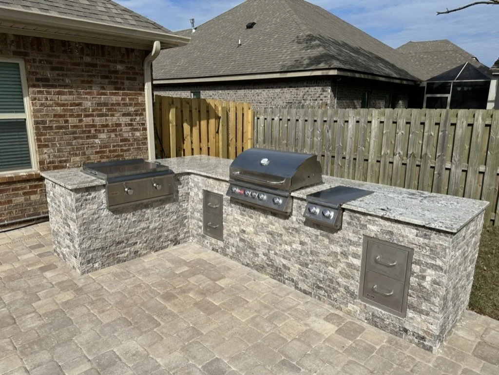 Outdoor kitchen with a stone countertop featuring a built-in grill, cooking station, and storage drawers on a paved patio, set against a brick wall and wooden fence.