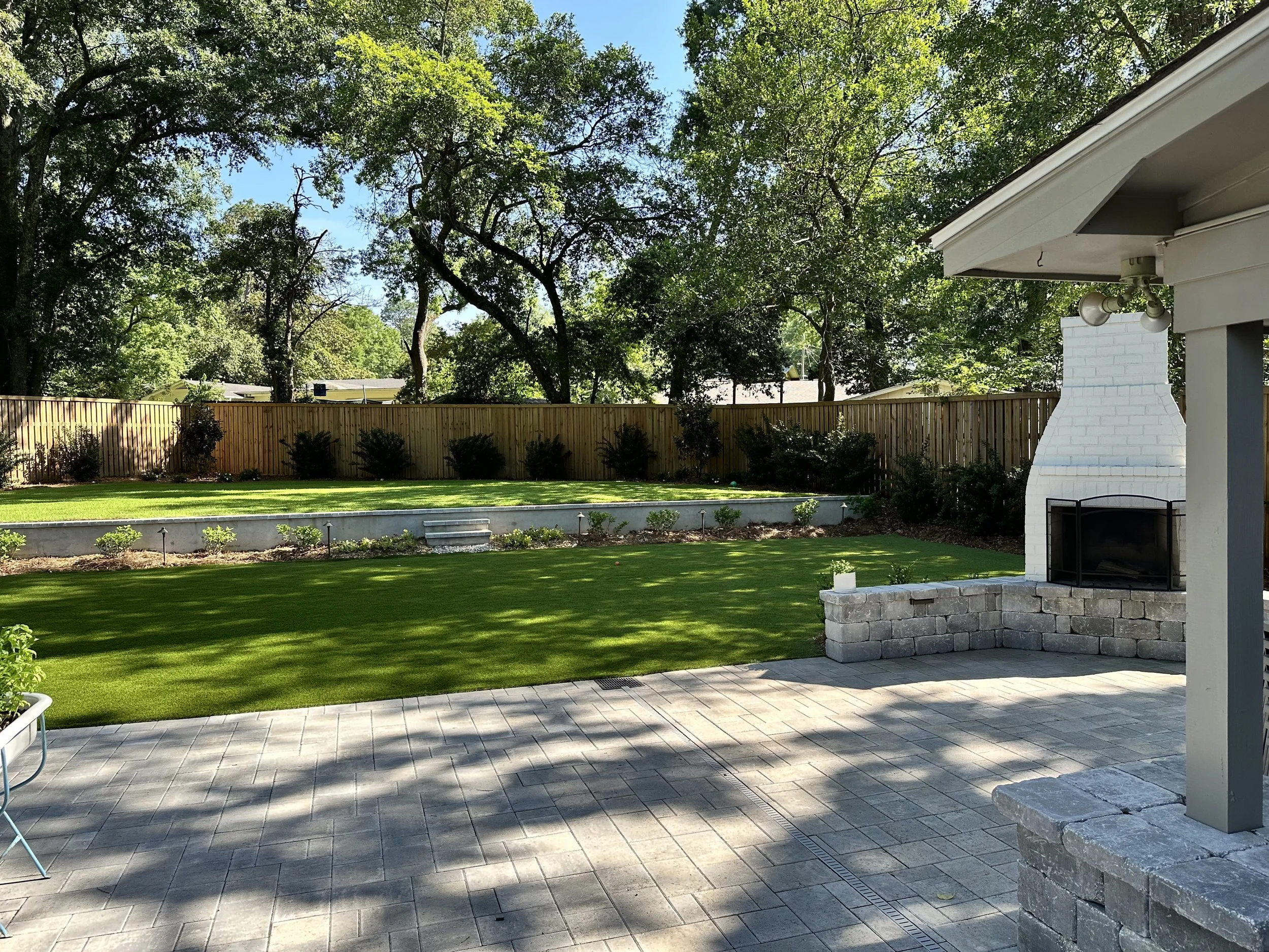 Beautiful Gulf Coast backyard with artificial turf, paver patio, and outdoor fireplace, surrounded by landscaped garden beds and trees.