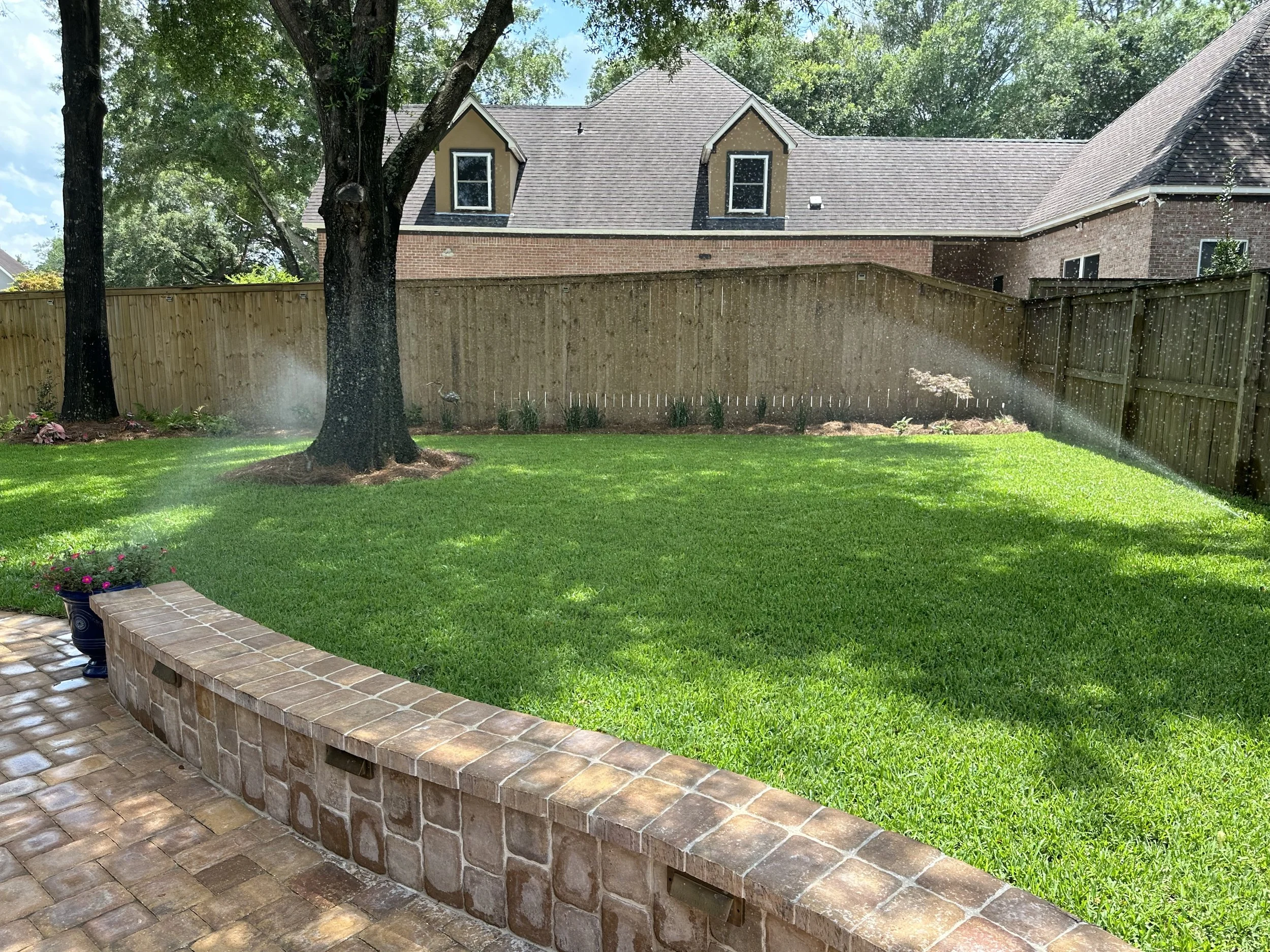 Backyard lawn with active irrigation system and brick retaining wall, providing consistent lawn care in a Gulf Coast setting.