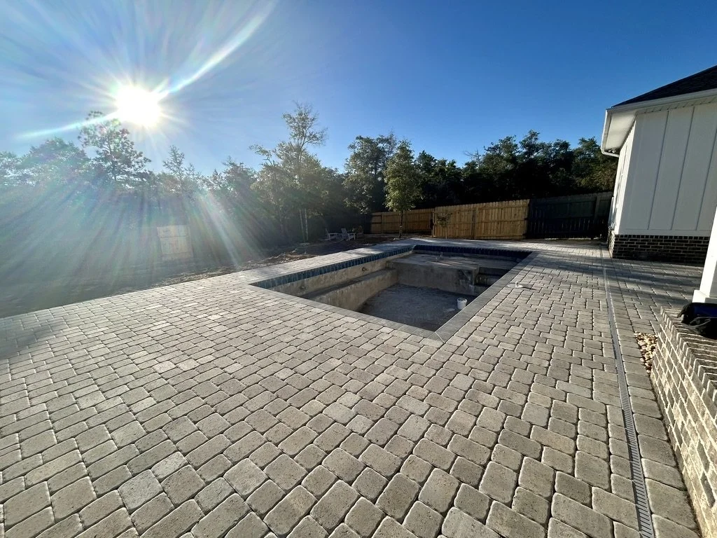 Sunlit backyard patio with stone paver flooring surrounding an unfinished in-ground pool, bordered by a wooden fence and surrounded by trees.