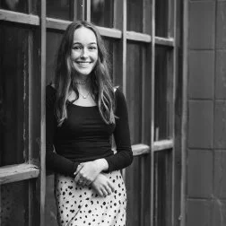 Young woman with long hair smiling, standing near a wooden structure or fence, in black and white.