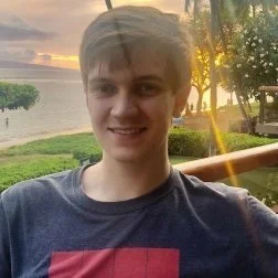Young man smiling outdoors near a body of water at sunset, with trees and a wooden fence in the background.