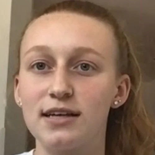 A young woman with red hair and earrings looking at the camera indoors.