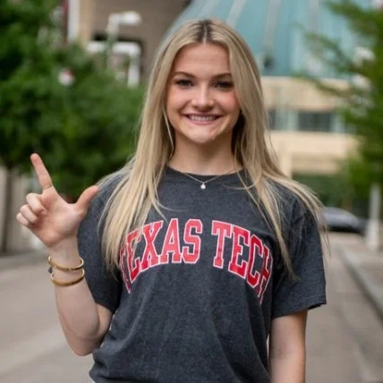 A young woman with long blonde hair smiling, wearing a Texas Tech t-shirt, standing outdoors.