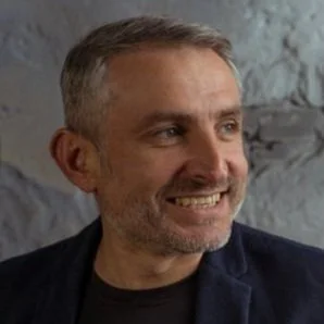 A man with short gray hair and a beard, smiling, wearing a black shirt and a dark jacket, standing against a gray wall.