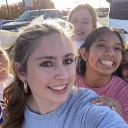 Four young women smiling outdoors, with cars and trees in the background.