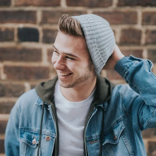 Young man smiling, wearing a gray knit beanie, denim jacket, and white shirt, standing in front of a brick wall.