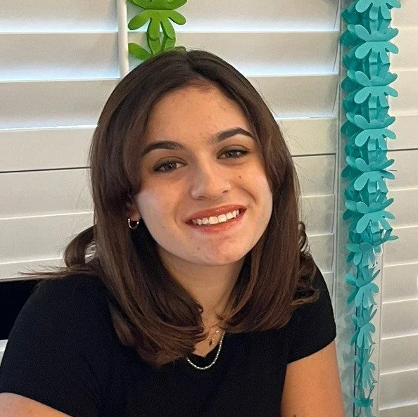 A young woman with shoulder-length brown hair, smiling and wearing a black top, earrings, a necklace, and a nose ring in front of white louvered walls decorated with teal paper flowers.