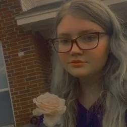 Young woman with long gray hair and glasses holding a cupcake with pink frosting, standing outside near a brick building.