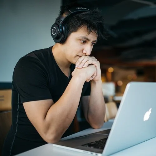 A man wearing headphones looks at a laptop screen with his hands clasped under his chin.