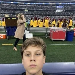 A young boy taking a selfie at a sports stadium during a game or event, with people in yellow jackets and team staff on the field in the background.