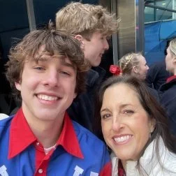 Two smiling people, a teenage boy with curly hair in a red and blue shirt, and a woman with dark hair and a red hair accessory, taking a selfie outdoors with a school bus and other children in the background.