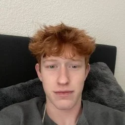 A young man with reddish hair taking a selfie in a room with a beige wall and black headboard.