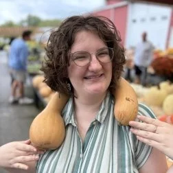 Woman with short curly hair and glasses smiling while holding two gourd-shaped objects on her shoulders at an outdoor event with people and farm produce in the background.