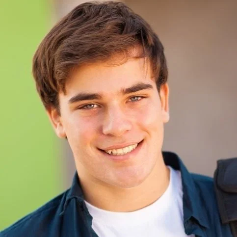 A young man with short brown hair and blue eyes smiling at the camera, wearing a dark jacket and white shirt, outdoors against a blurred background.