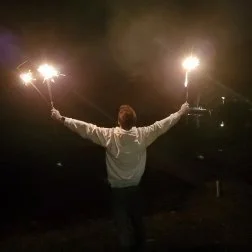 Person holding two sparklers at night with arms extended