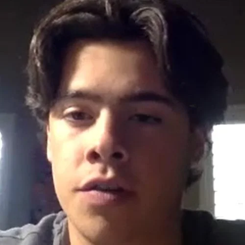 Close-up of a young man with dark wavy hair looking at the camera in a dimly lit room.