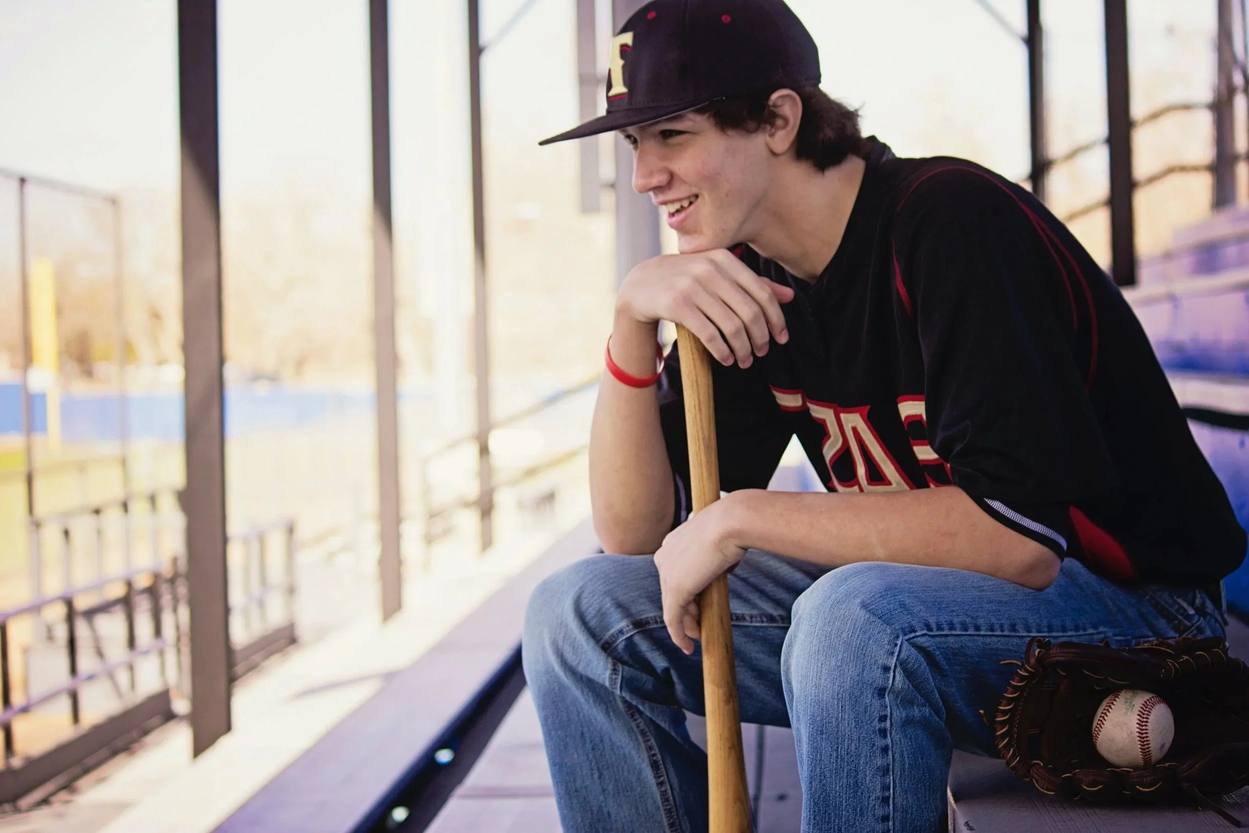 A teenage boy sitting on baseball dugout bleachers holding a baseball bat with a baseball and glove nearby, smiling, wearing a dark sports jersey and cap.