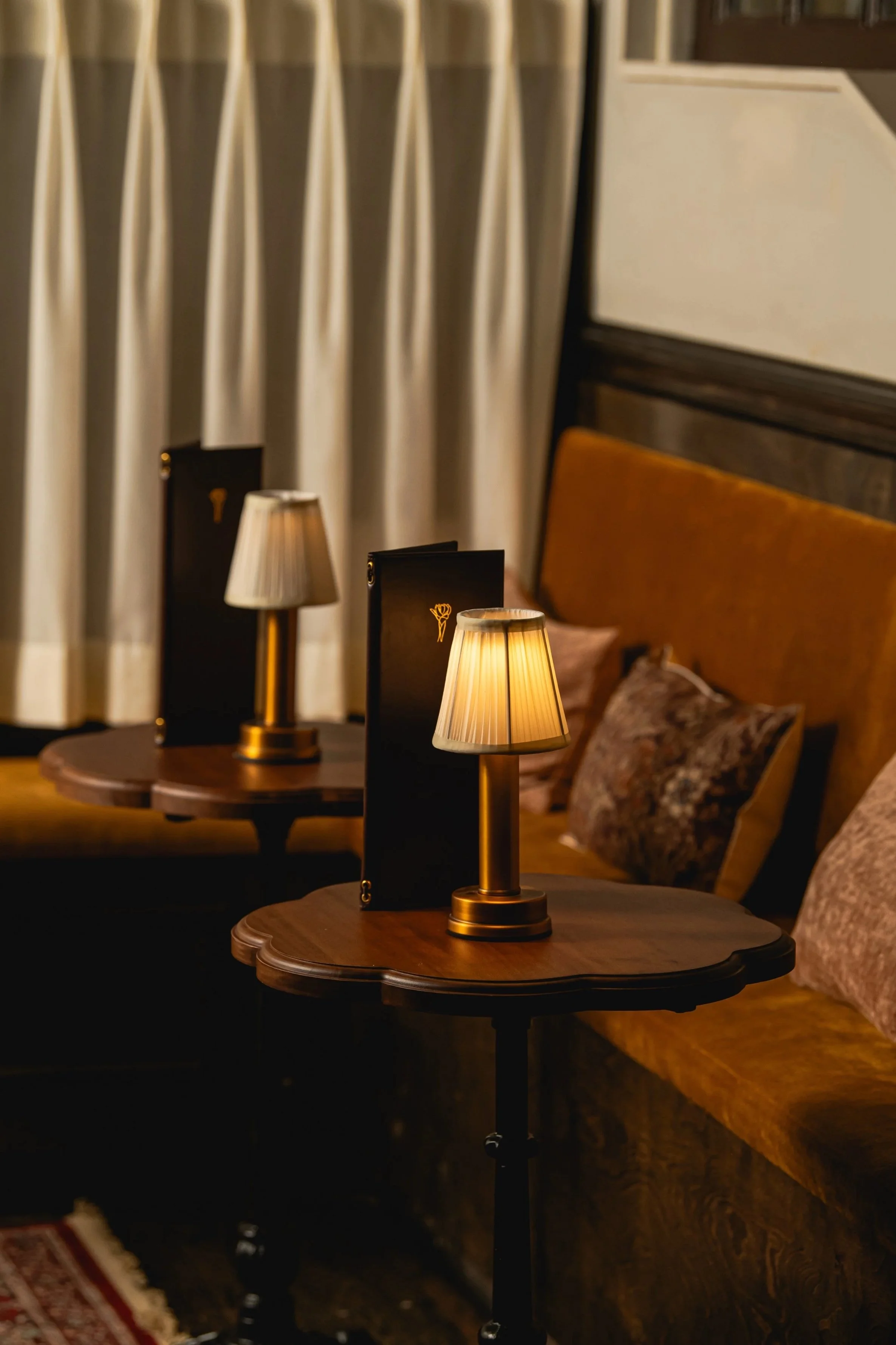 A cozy indoor setting with two small wooden tables, each holding a lit table lamp with beige shades, black menus, daytime curtains, a plush sofa with pillows, and warm ambient lighting.
