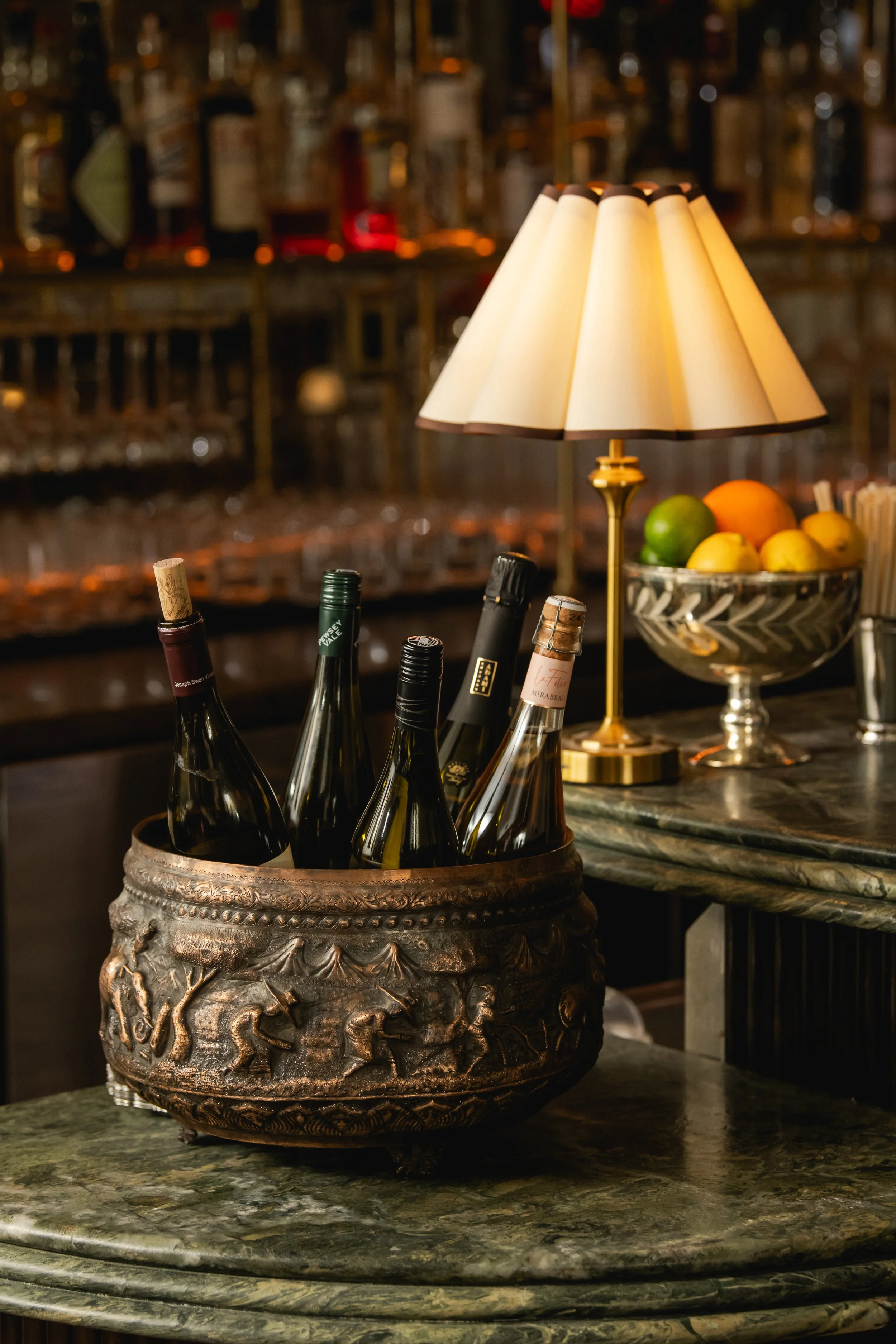 A decorative metal bowl filled with wine bottles on a marble countertop, a table lamp, and a glass bowl of assorted fruits on a bar counter.