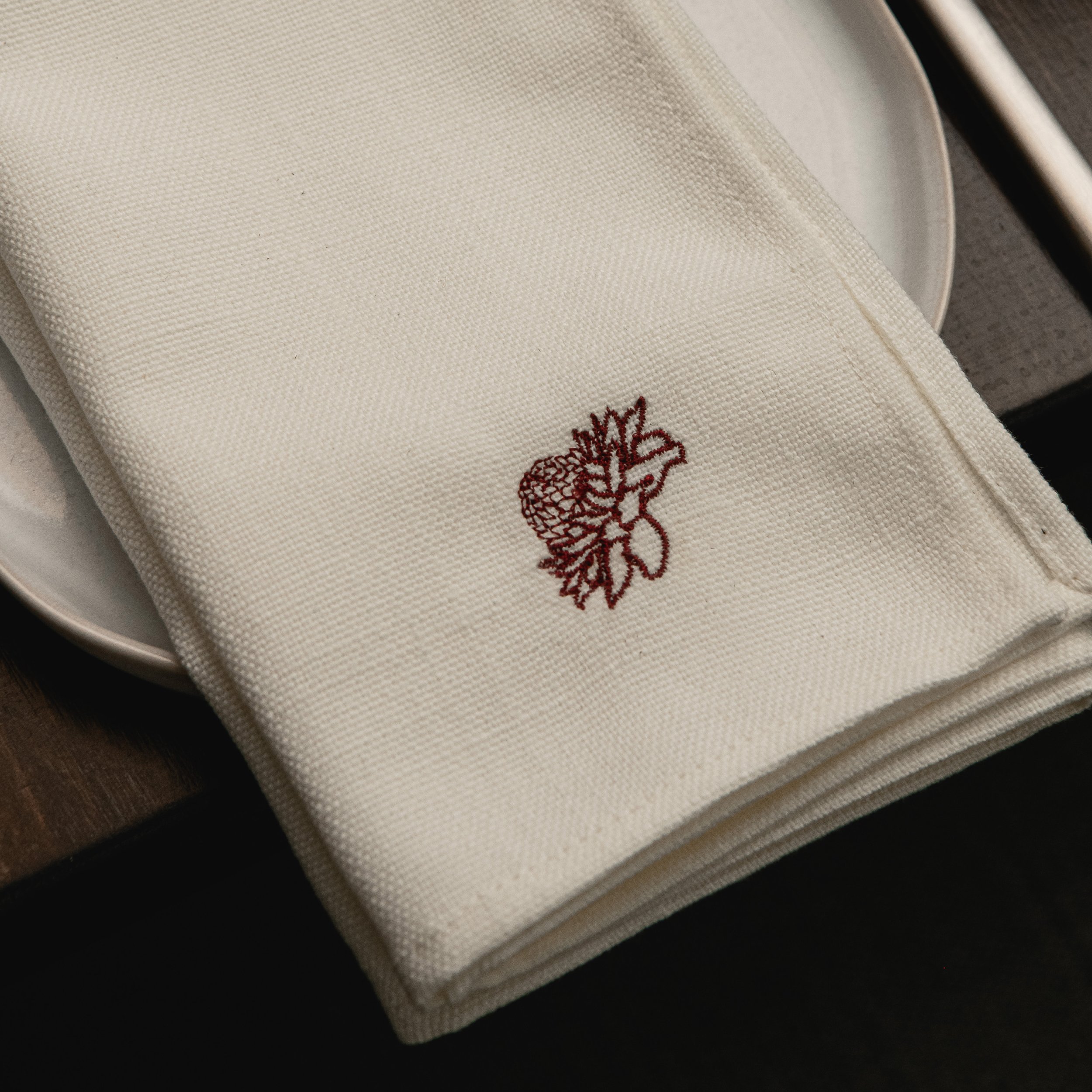 A folded white cloth napkin with a red embroidered floral design, placed on a white plate.