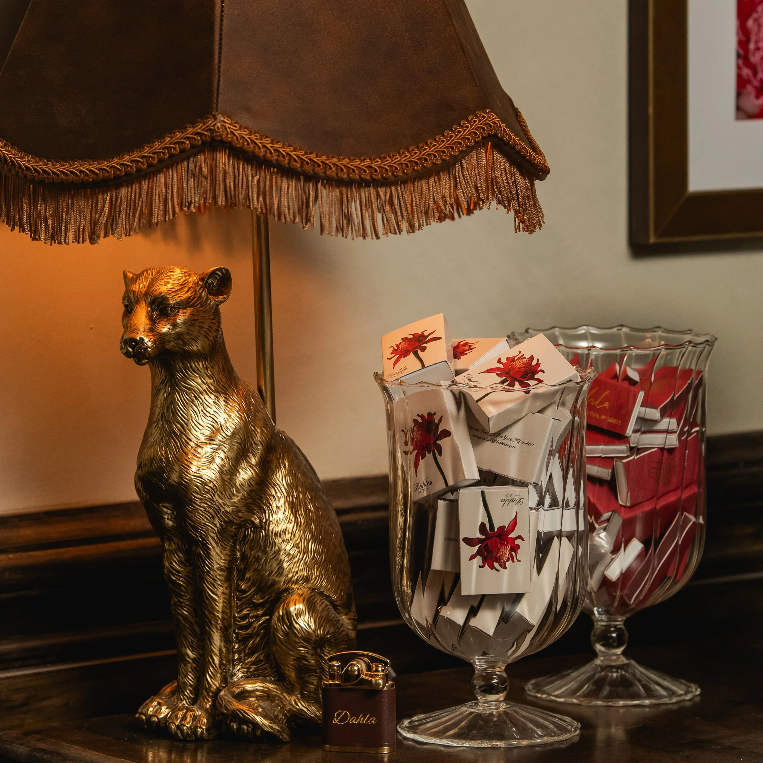 A gold-colored lion statue sitting on a wooden surface next to a glass lamp with a fringed brown lampshade, and two glass containers filled with small boxes, one with white and red packaging and the other with mostly red packaging. A small brown lighter with the word 'Dahla' is also present.