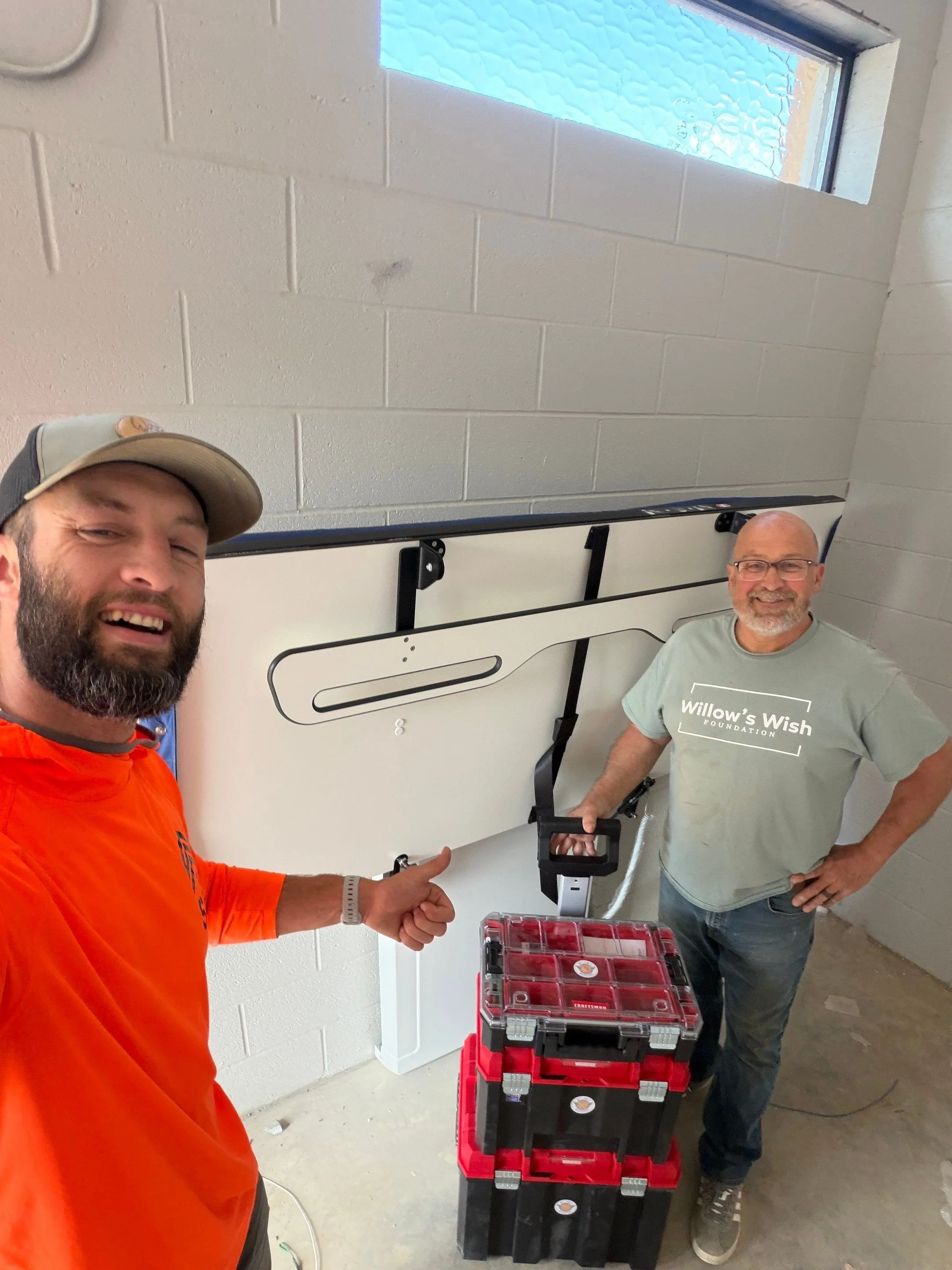 Mark Dail and a Willow's Warrior volunteer standing in a room with a newly installed powered changing table, white brick walls, with various red and black toolboxes on a table in front of them.