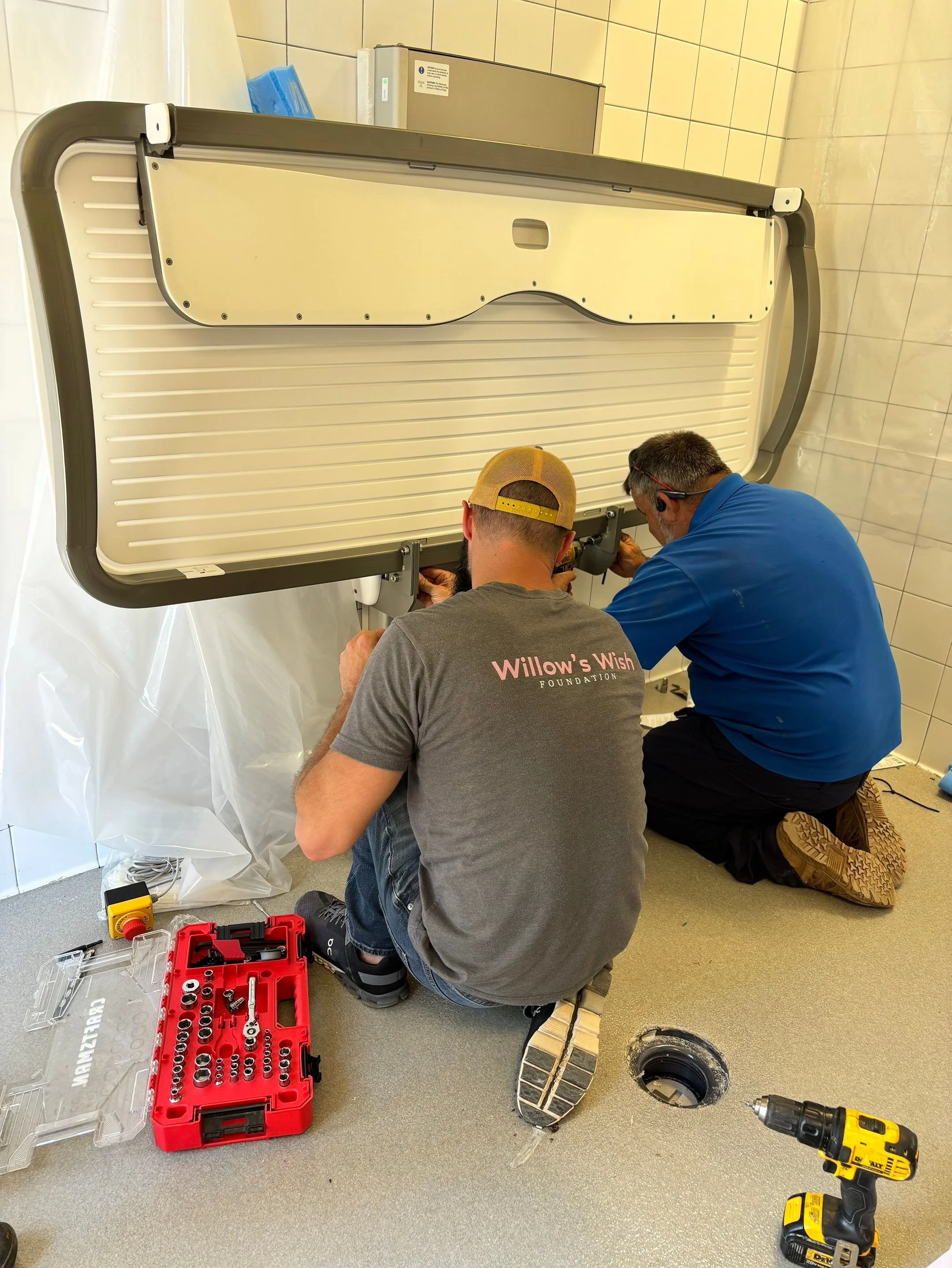 Two men working on installing a powered changing table in a tiled room. One is kneeling while the other is sitting, with various tools and equipment around them.