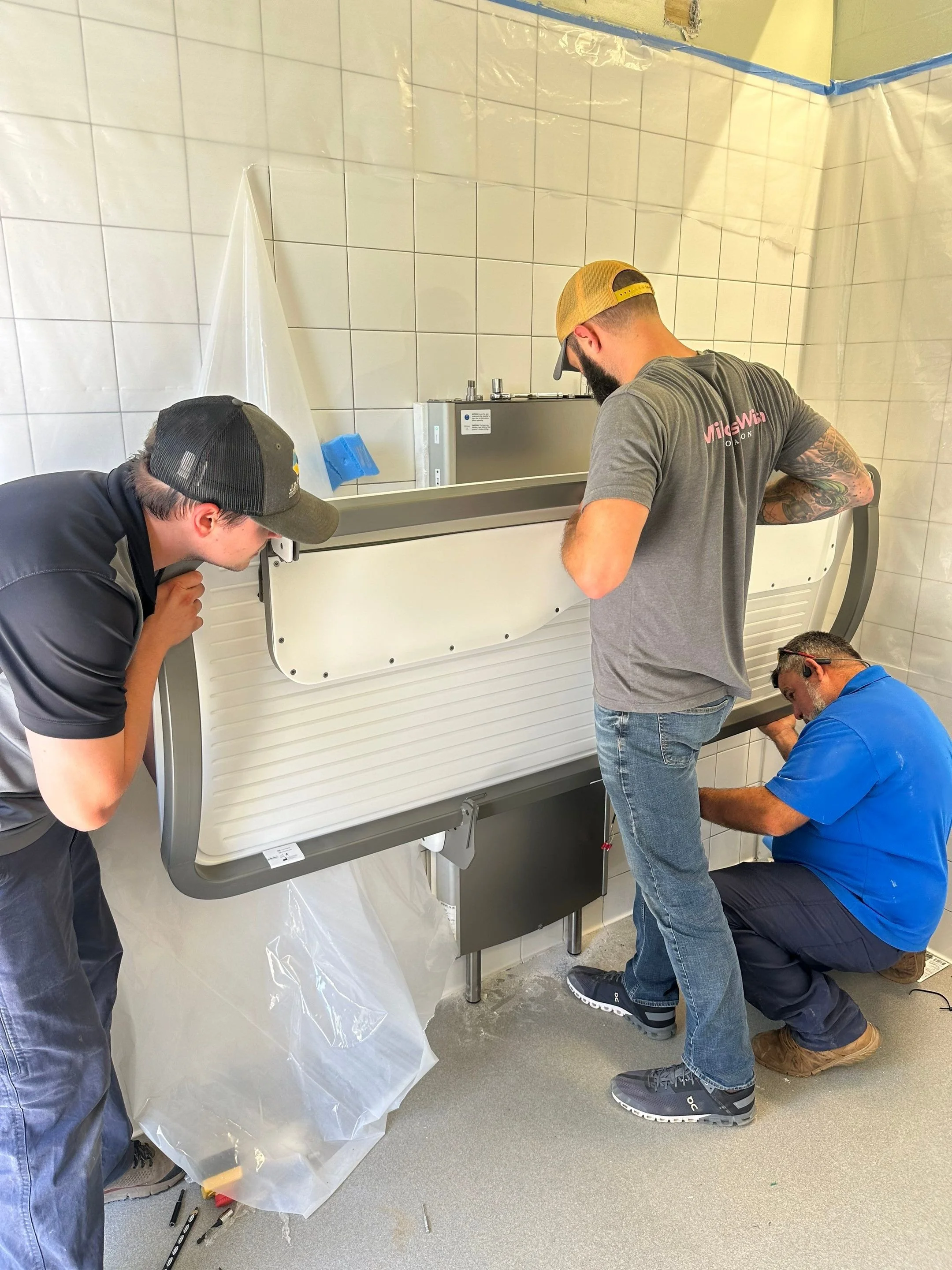 Willow's Warriors working together to install or repair a large white appliance, possibly a commercial refrigerator or freezer, in a room with tiled walls.