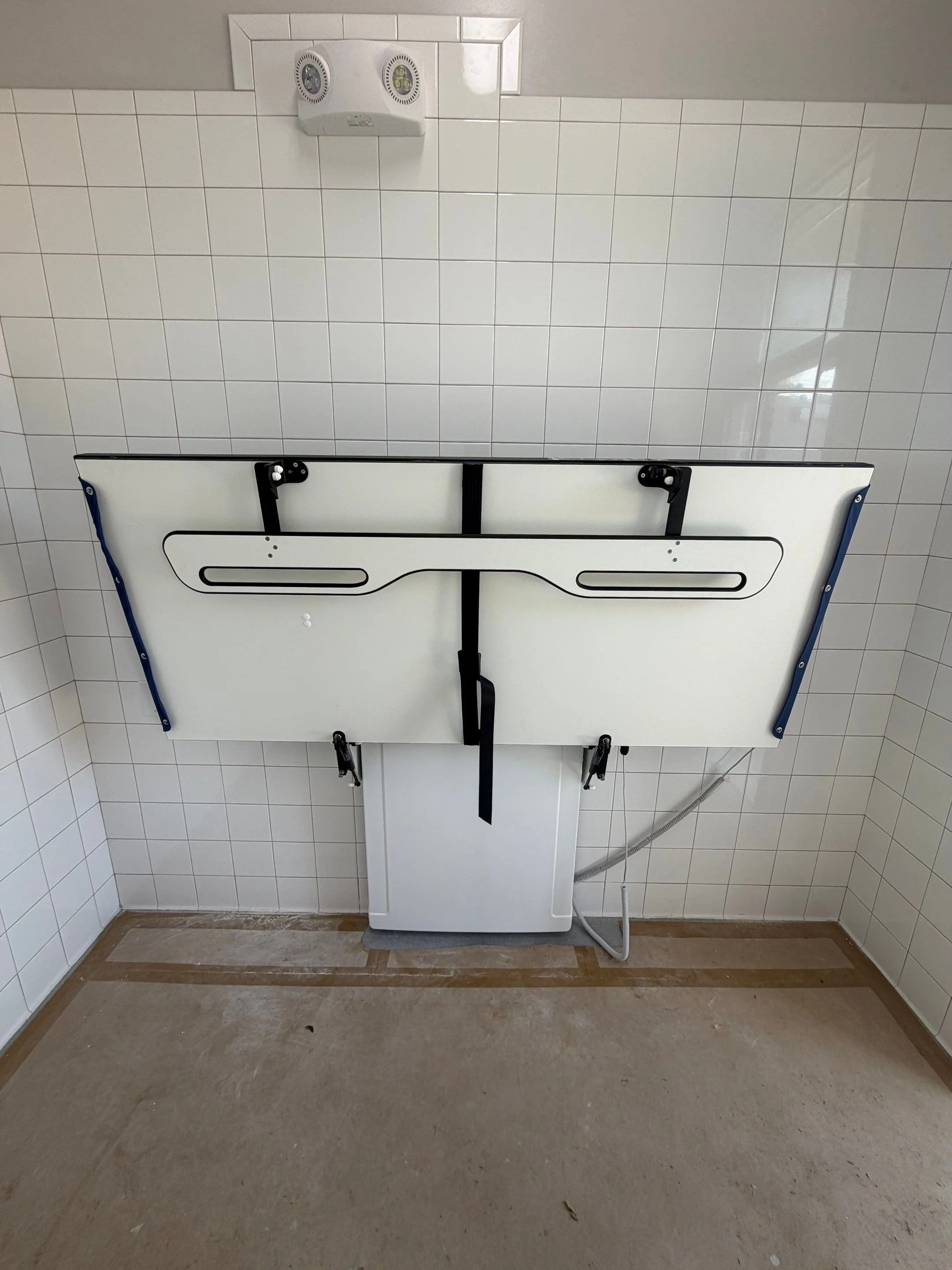 A white wall-mounted baby changing station with black straps and a flap, folded down in a room with white tiled walls and a gray tile ceiling. There is a power outlet and a white exhaust fan on the ceiling.