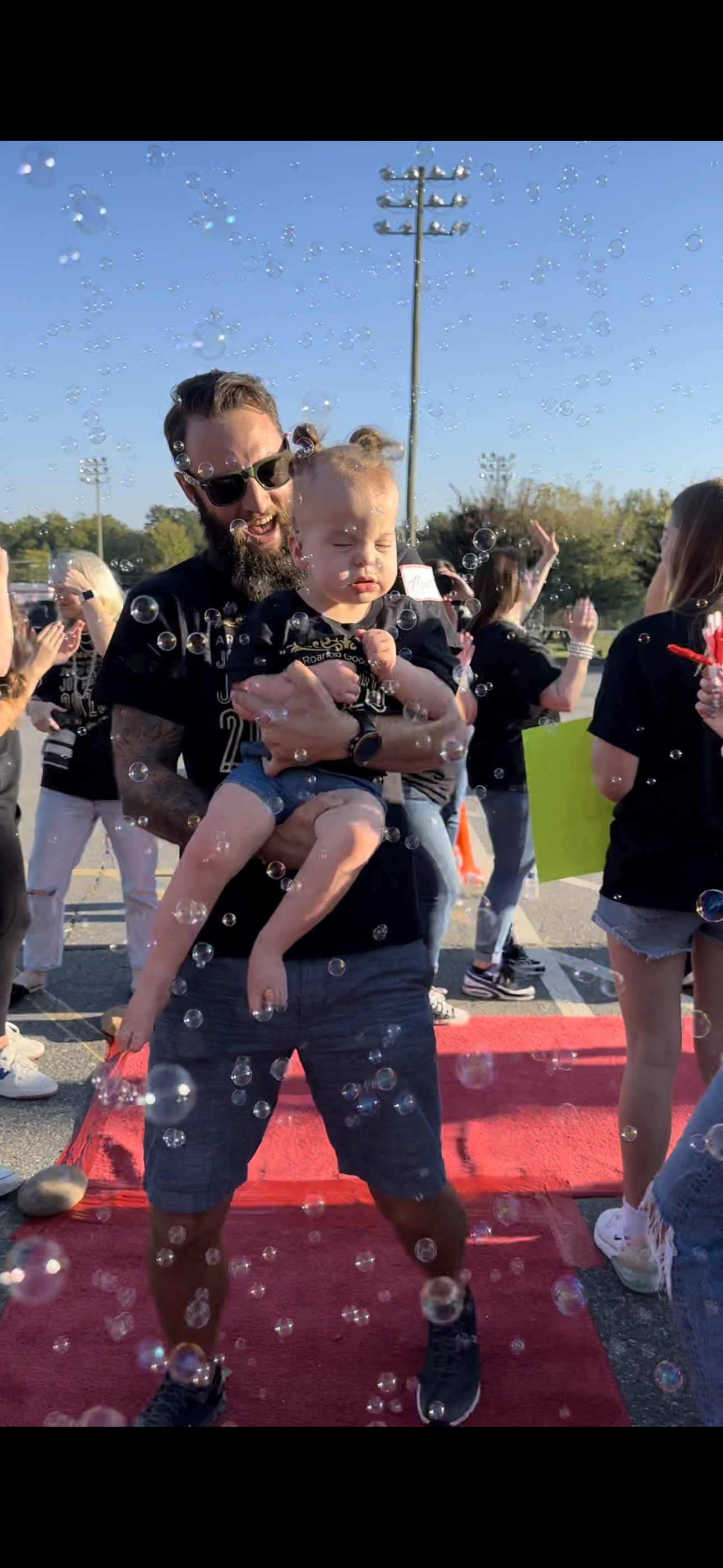 Mark Dail with a beard and sunglasses holding a Willow Dail surrounded by people, bubbles, and a red carpet, outdoors on a sunny day.