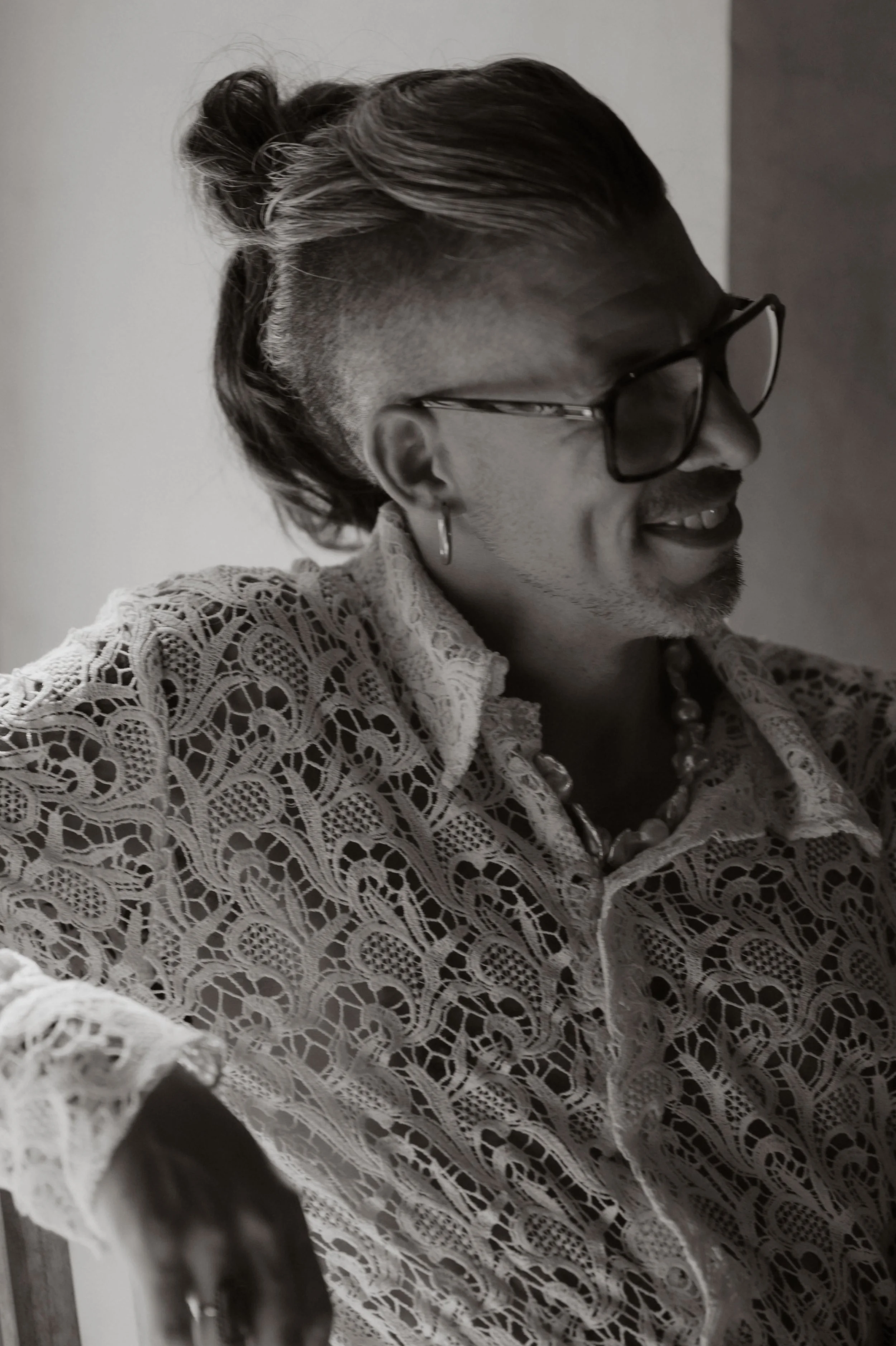 A man with glasses, earrings, and a lace shirt smiling and looking to the side.