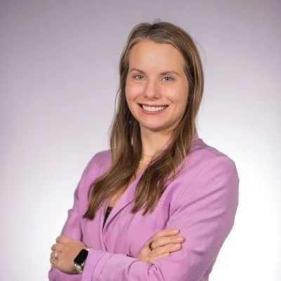 A woman with long, light brown hair smiling wearing a purple blazer and standing against a neutral background.