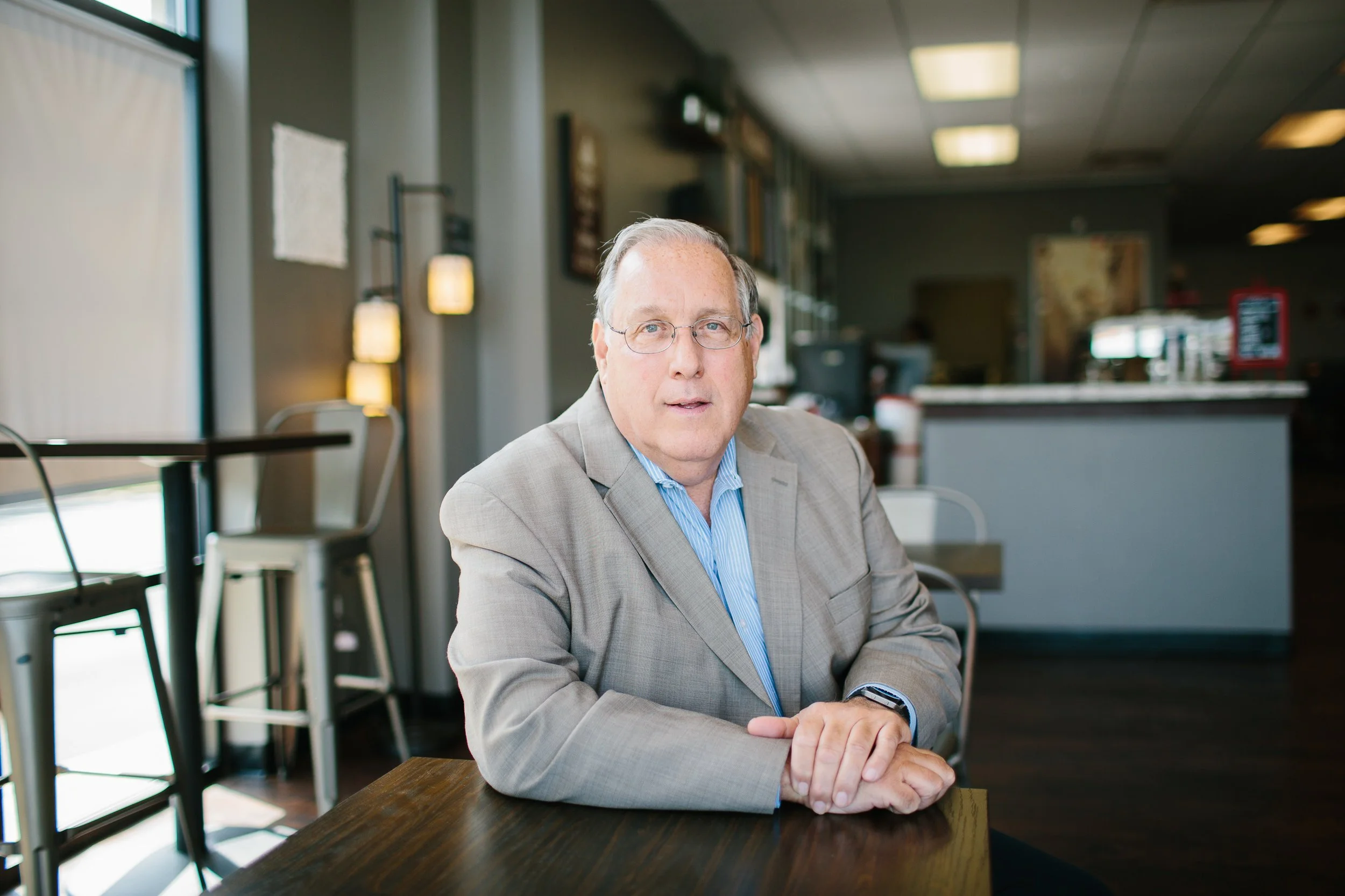 A middle-aged man in a beige suit and blue shirt sitting at a wooden table in a cafe with natural light coming through the windows.