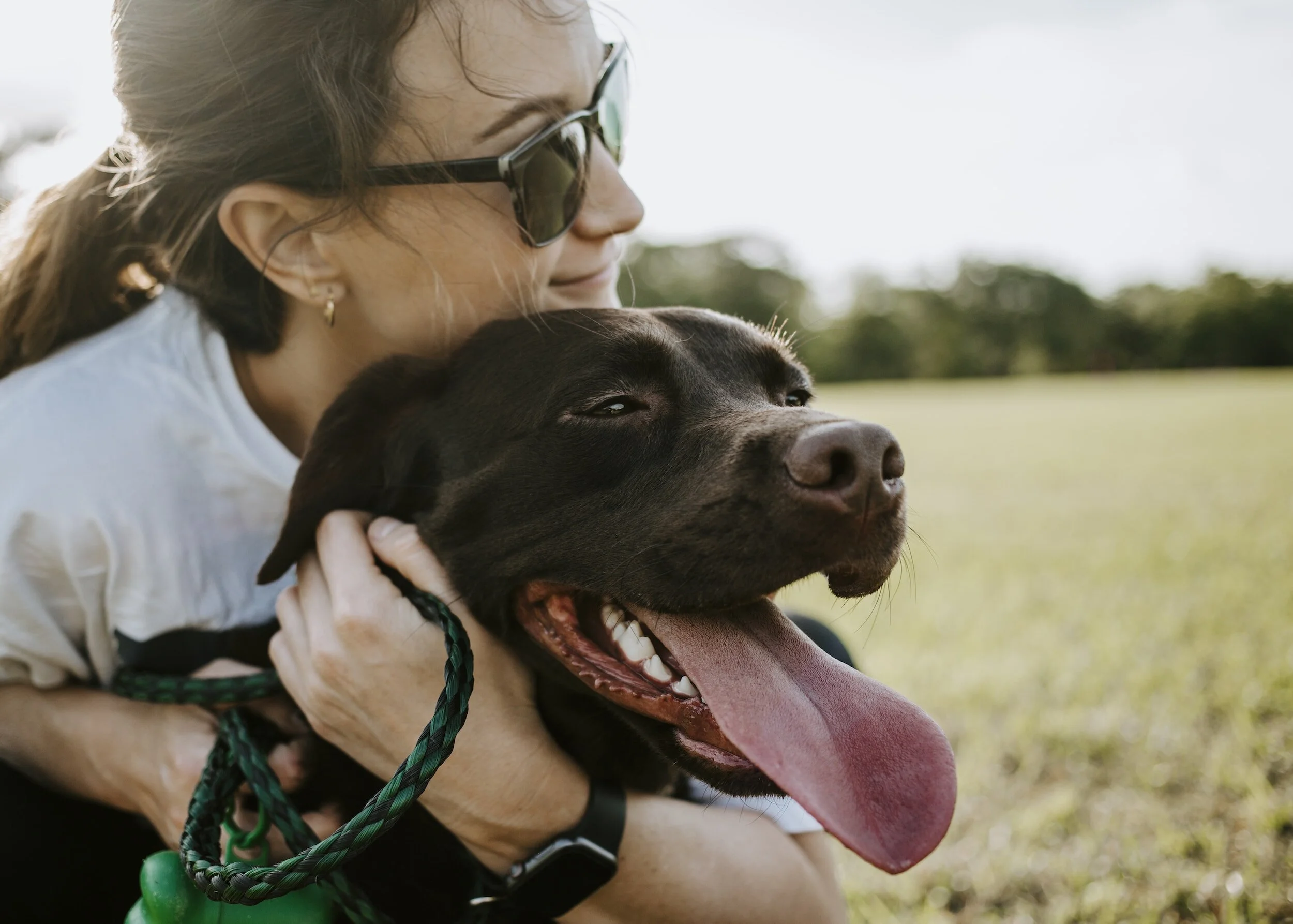 A woman wearing sunglasses and earrings hugs a happy black dog outdoors on a grassy field. The dog has its tongue out and appears content.