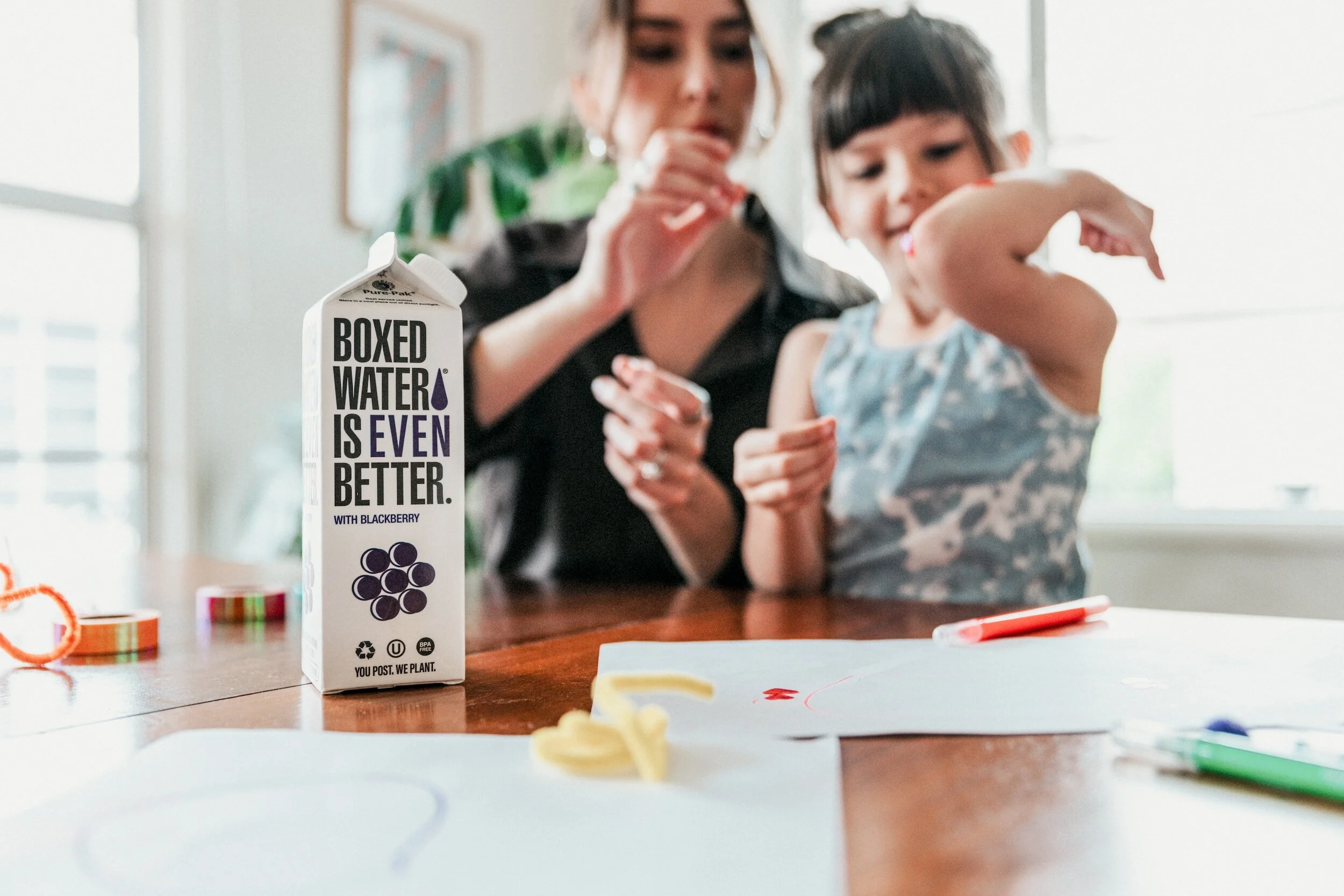 A woman and a young girl are sitting at a table, with the woman drinking from a box of blackberry-flavored water. The table has papers, markers, and rubber bands on it.