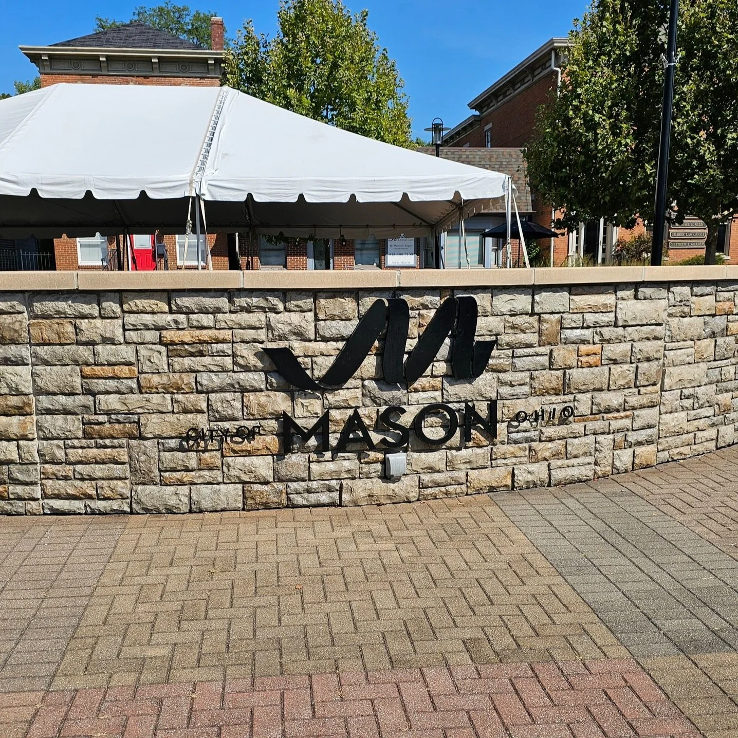 Stone brick wall with a sign that reads 'City of Mason' with the year 2010, and a large black 'M' logo above the text. In the background, there is a white tent and brick buildings with trees and a blue sky.