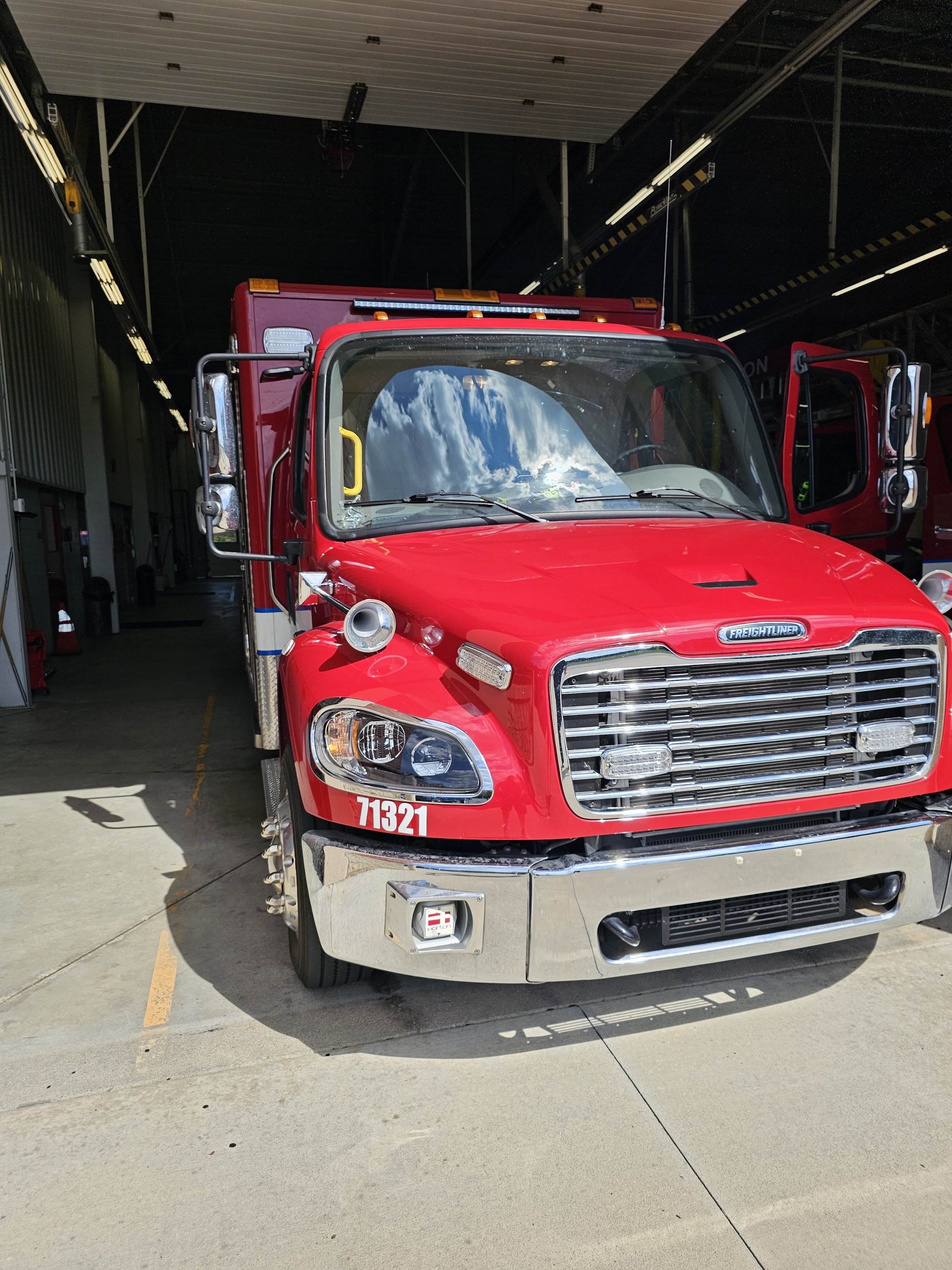 Red fire truck parked inside a fire station with blue sky and clouds reflected in the windshield.