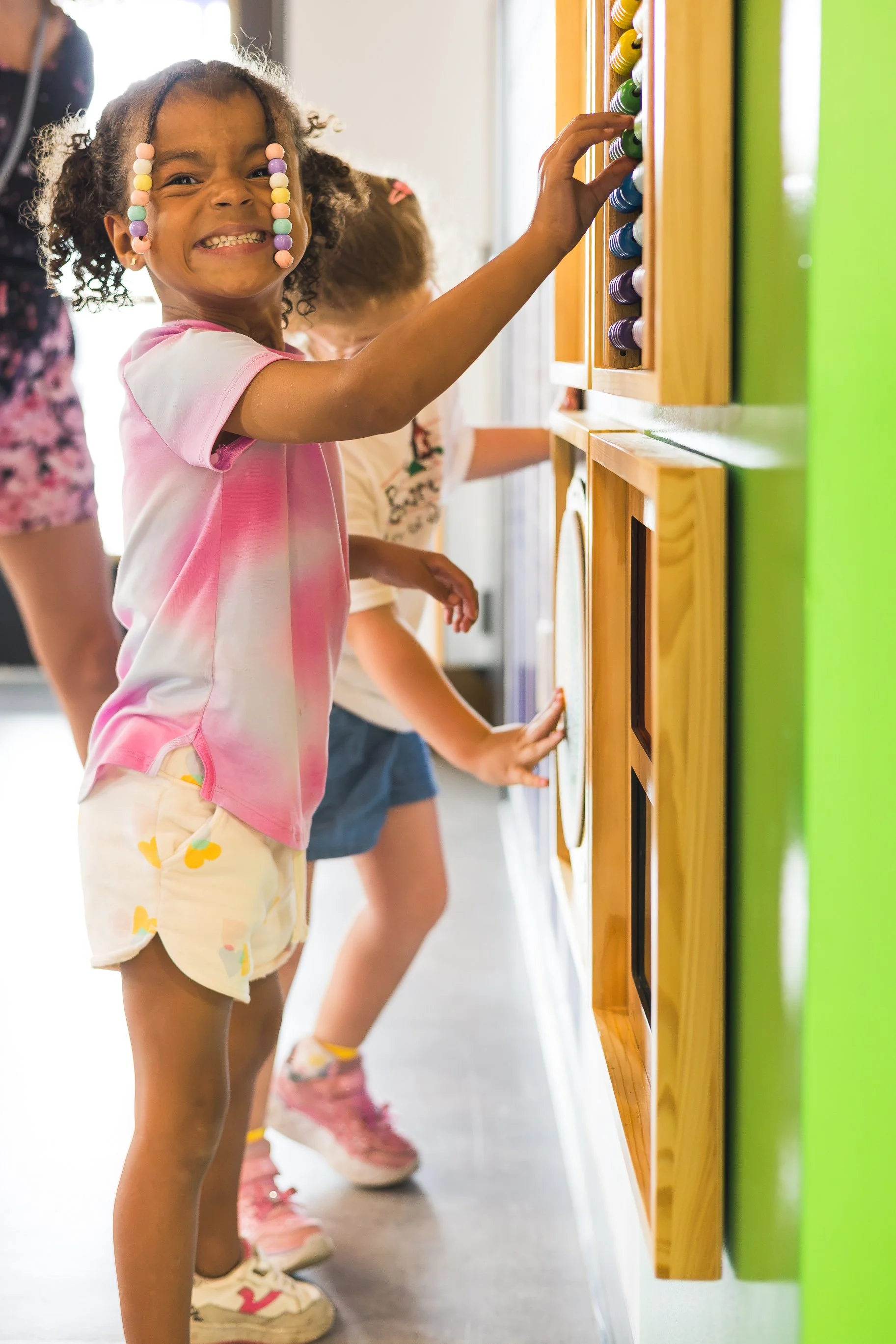 A child smiles at the camera while playing with a sensory board on the wall