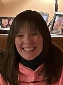 Smiling woman with dark hair and bangs, wearing a pink top, in front of a wall with framed photos.