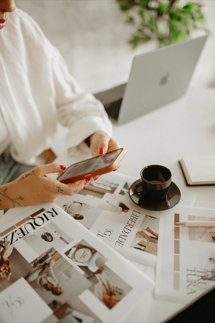 A person with red nail polish and tattoos on their hand is using a smartphone on a desk with fashion magazines and a black coffee cup. A laptop is open nearby and a notebook with a pen is also on the desk.