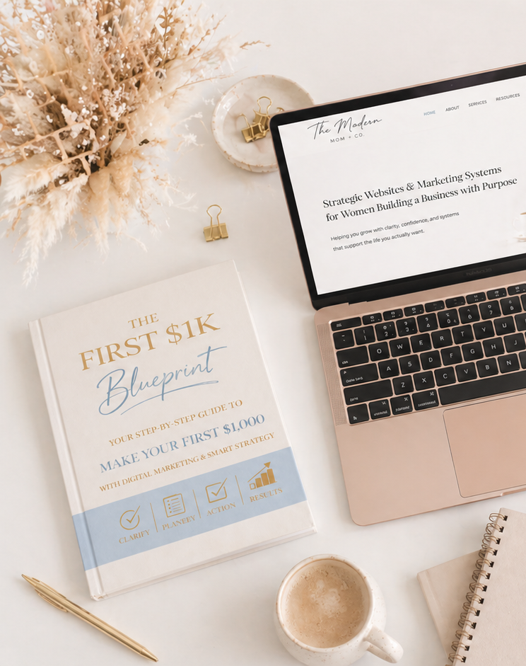 A flat lay photo of a workspace featuring a book titled 'The First $1K Blueprint', a partially open laptop displaying a website about marketing systems for women, a white cup of coffee, a gold pen, a notebook, a small dish with gold paper clips, and a vase with dried flowers on a white surface.