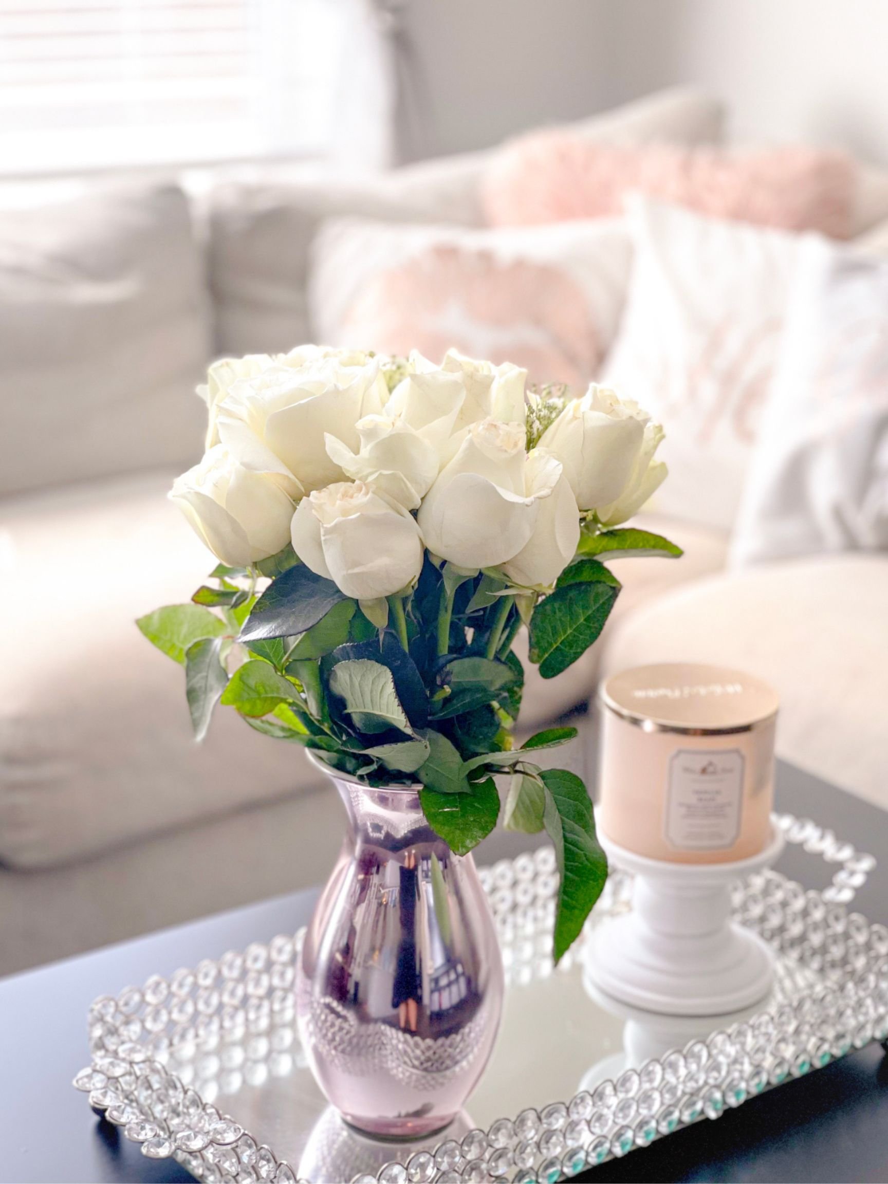 A bouquet of white roses in a pink metallic vase on a silver tray, with a lit candle in a white holder nearby, placed on a black table in a cozy, softly lit living room. The Modern Mom + Co Lifestyle decor. Selkirk Manitoba Canada. Amazon Storefront