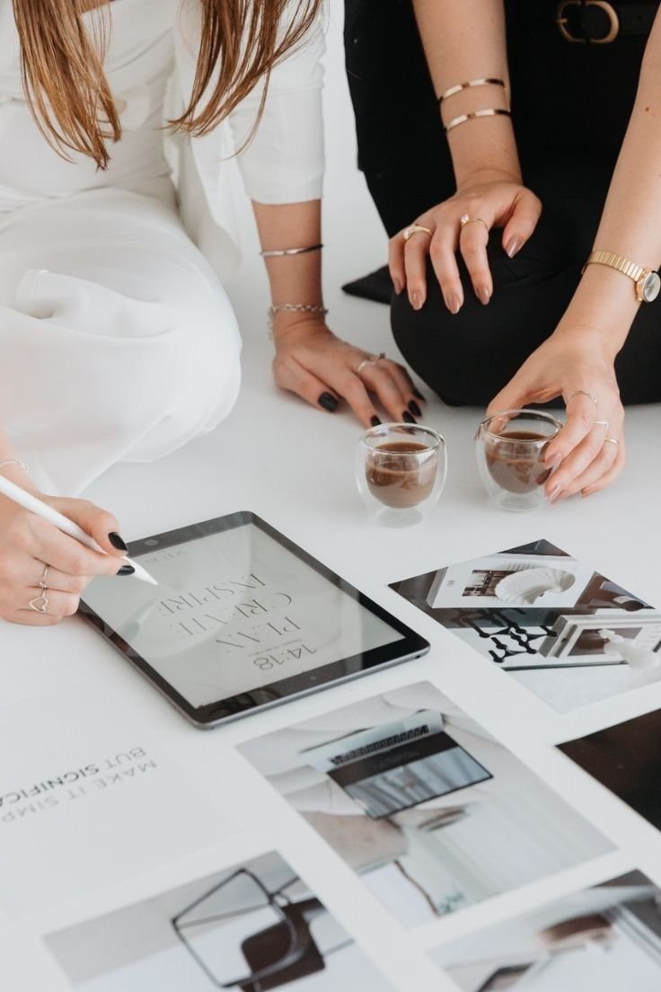 Two women are sitting at a white table, one holding a tablet with text and the other lifting glasses of coffee. The table is decorated with photos and printed material, and one woman is writing on the tablet with a stylus.