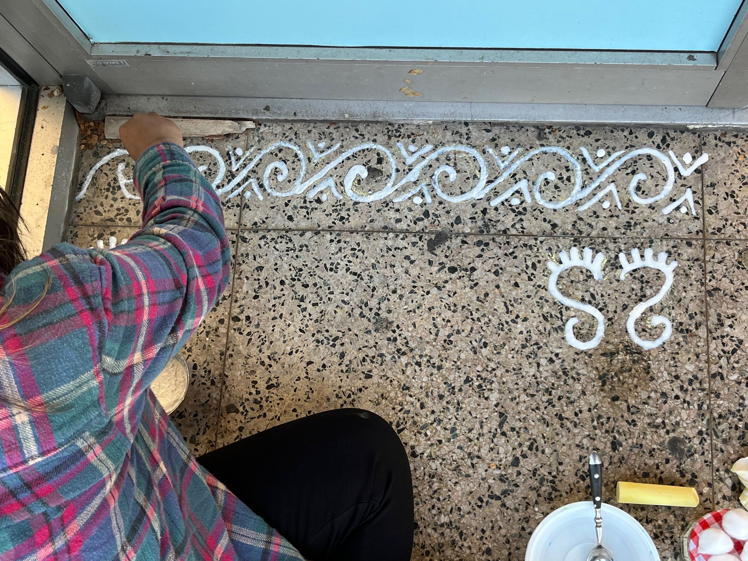 Artist Tanya Gupta makes an alpona, a traditional design from the Bengali culture of India, using powdered rice flour and water. The design, done at the threshold, includes a simple depiction of the feet of goddess Lakshmi, welcoming her to enter the