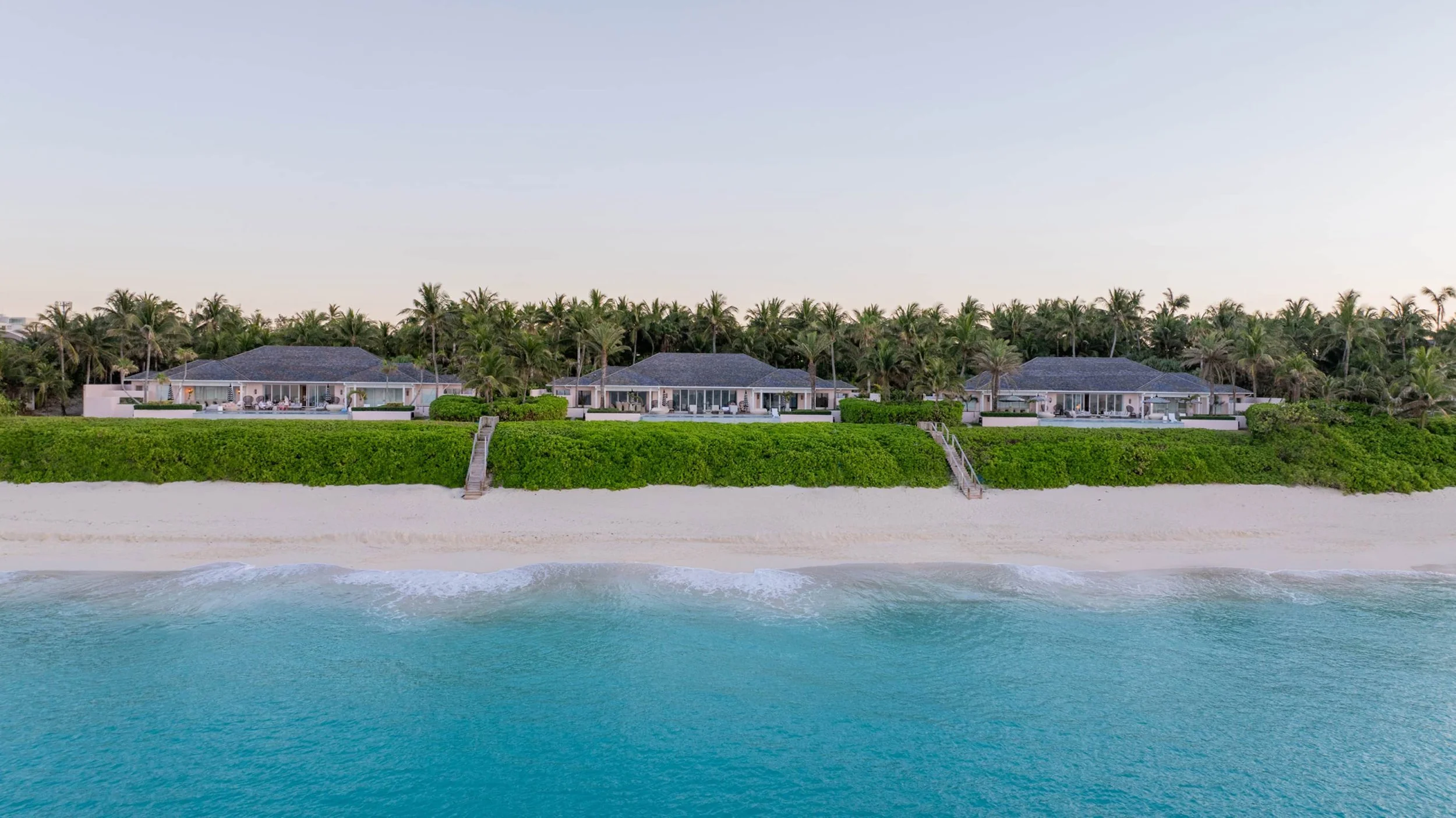 Beachfront luxury homes with stairs leading to sandy beach, lush greenery, palm trees, and calm ocean waves under a clear sky.