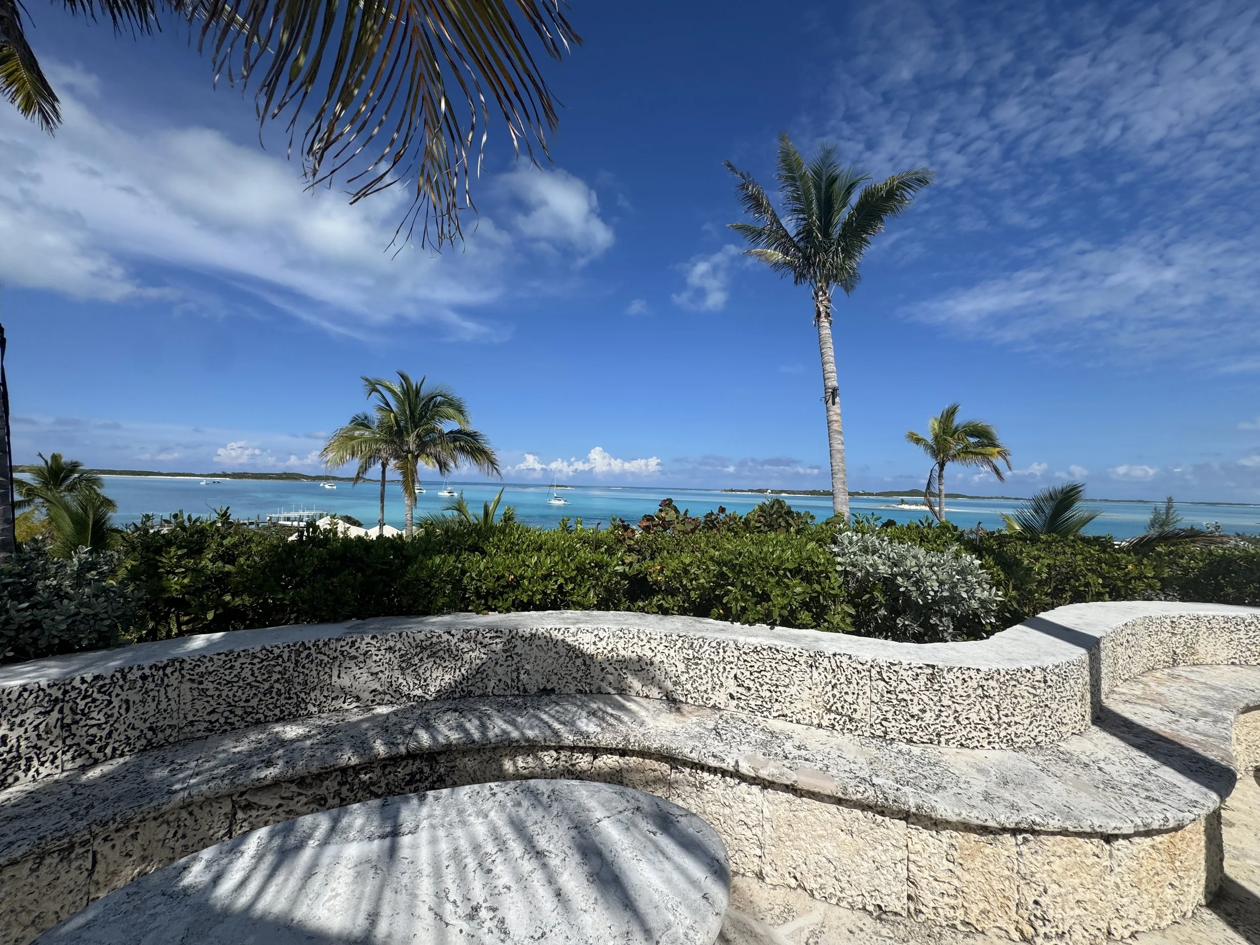View of a beach with palm trees, blue water, and sailboats under a partly cloudy sky.