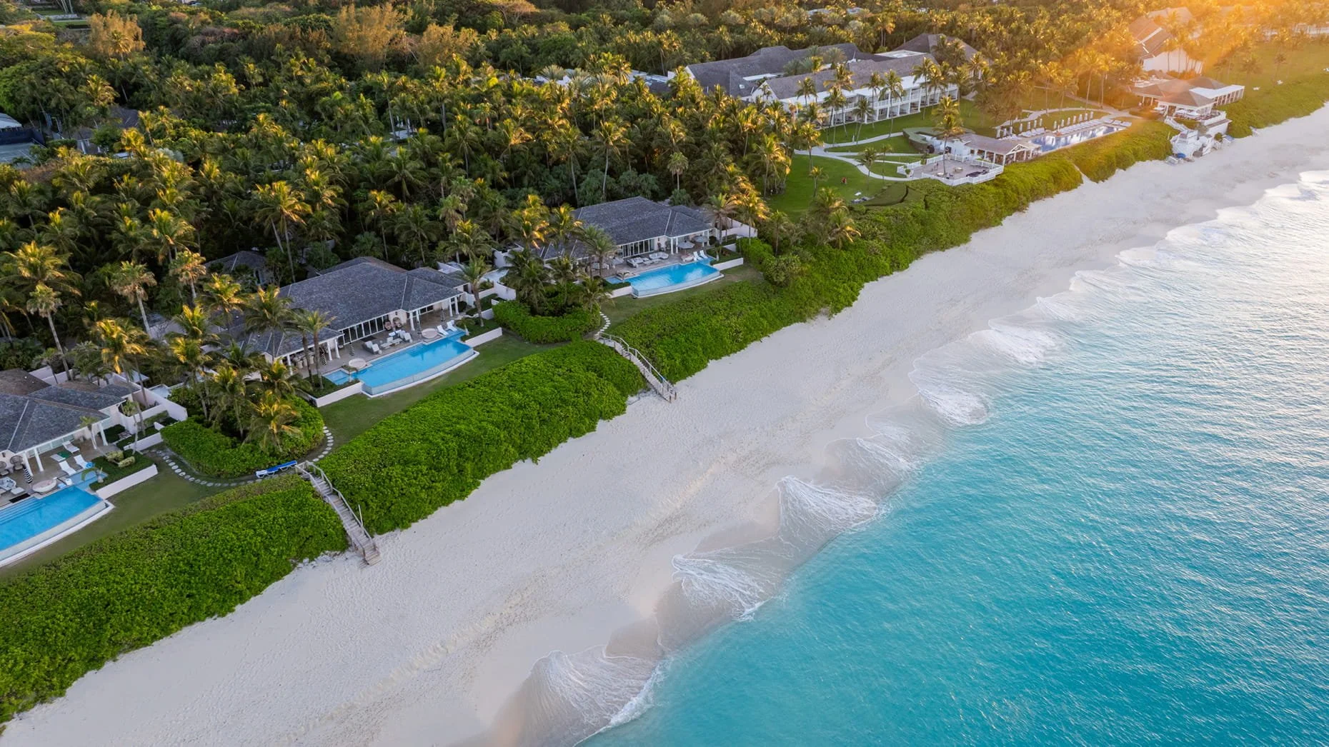Aerial view of beachfront resort with multiple villas, each with private pools, surrounded by palm trees and lush greenery, bordered by white sandy beach and turquoise ocean.